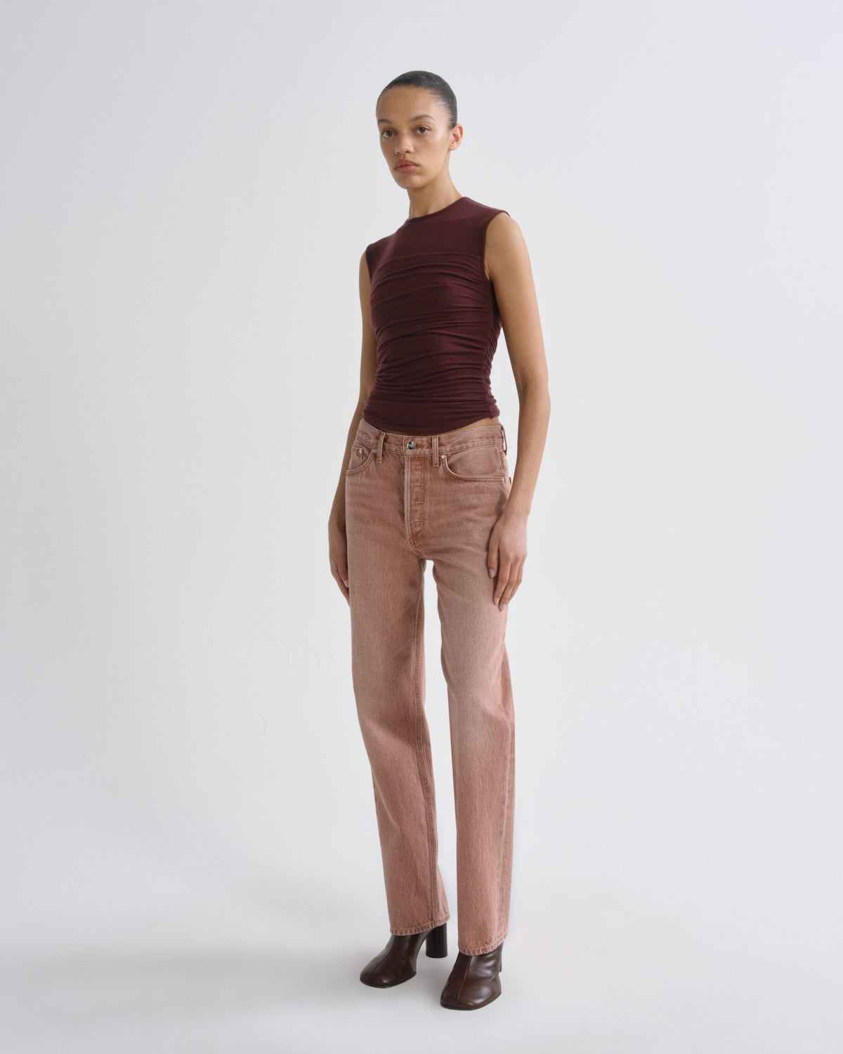 A model stands against a light gray studio background wearing a sleeveless dark burgundy top with gathered detailing and straight-leg dusty rose pants with dark brown heeled boots.