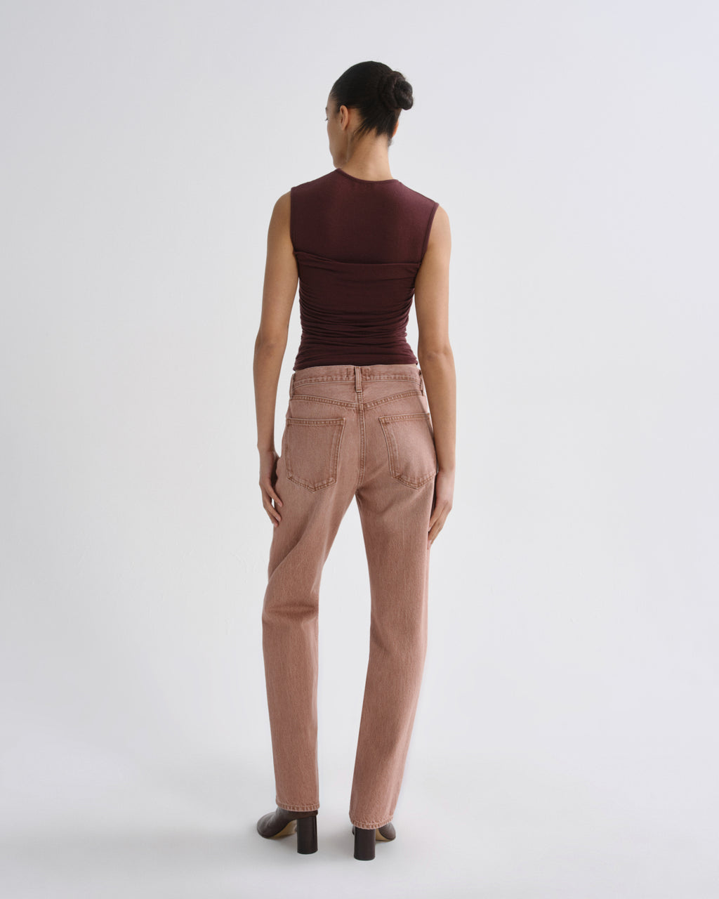 A model shown from the back wearing a sleeveless dark burgundy top and dusty rose straight-leg jeans, standing against a plain light gray studio background.