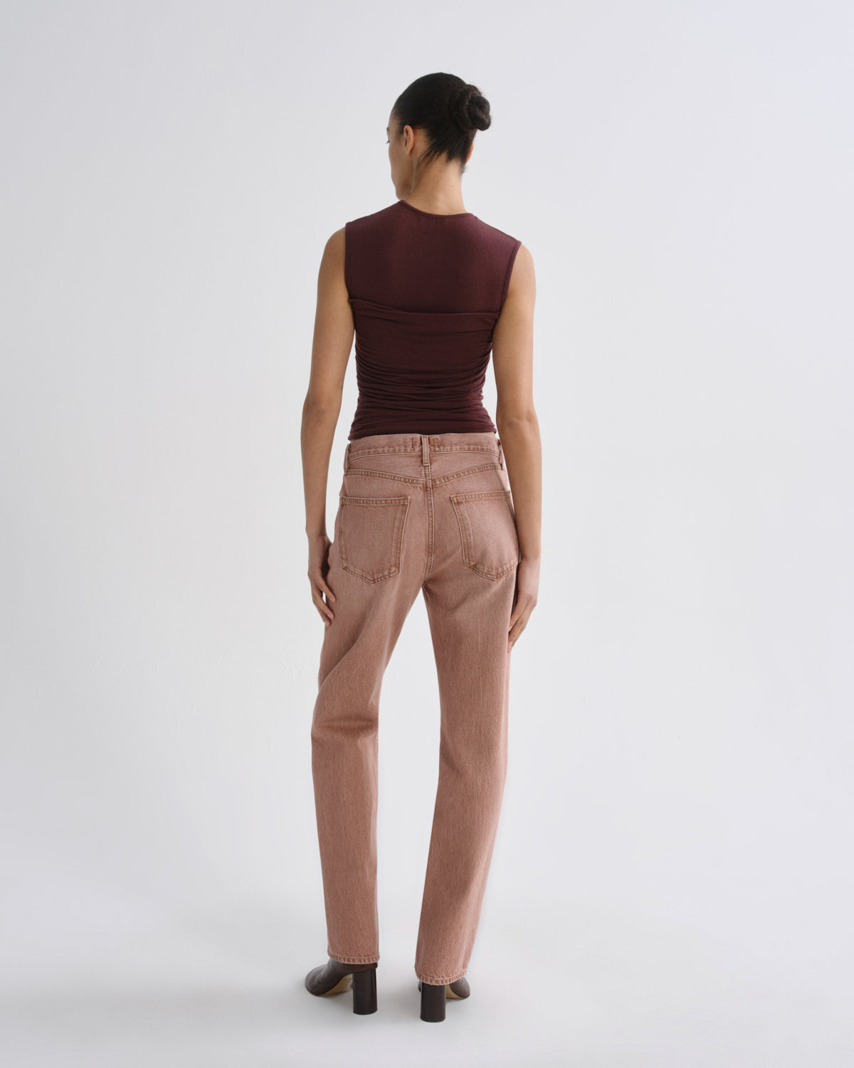 A model shown from the back wearing a sleeveless dark burgundy top and dusty rose straight-leg jeans, standing against a plain light gray studio background.