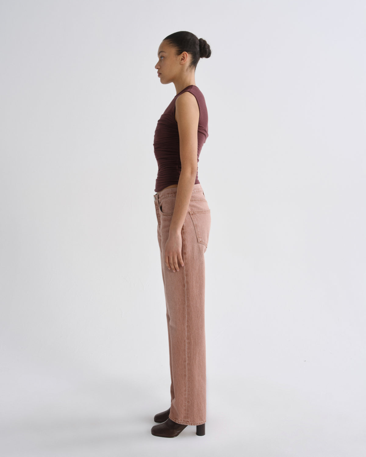 A woman stands in profile wearing a dark burgundy sleeveless top, dusty pink wide-leg jeans, and dark brown heeled shoes against a plain light gray studio background.