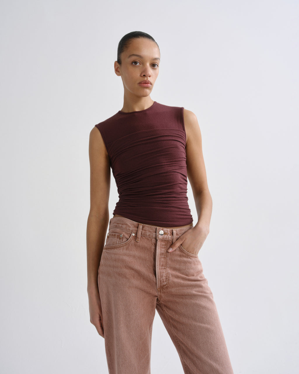A model wearing a sleeveless, ruched dark burgundy top and light brown high-waisted jeans, standing against a light gray studio background.