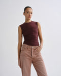 A model wearing a sleeveless, ruched dark burgundy top and light brown high-waisted jeans, standing against a light gray studio background.