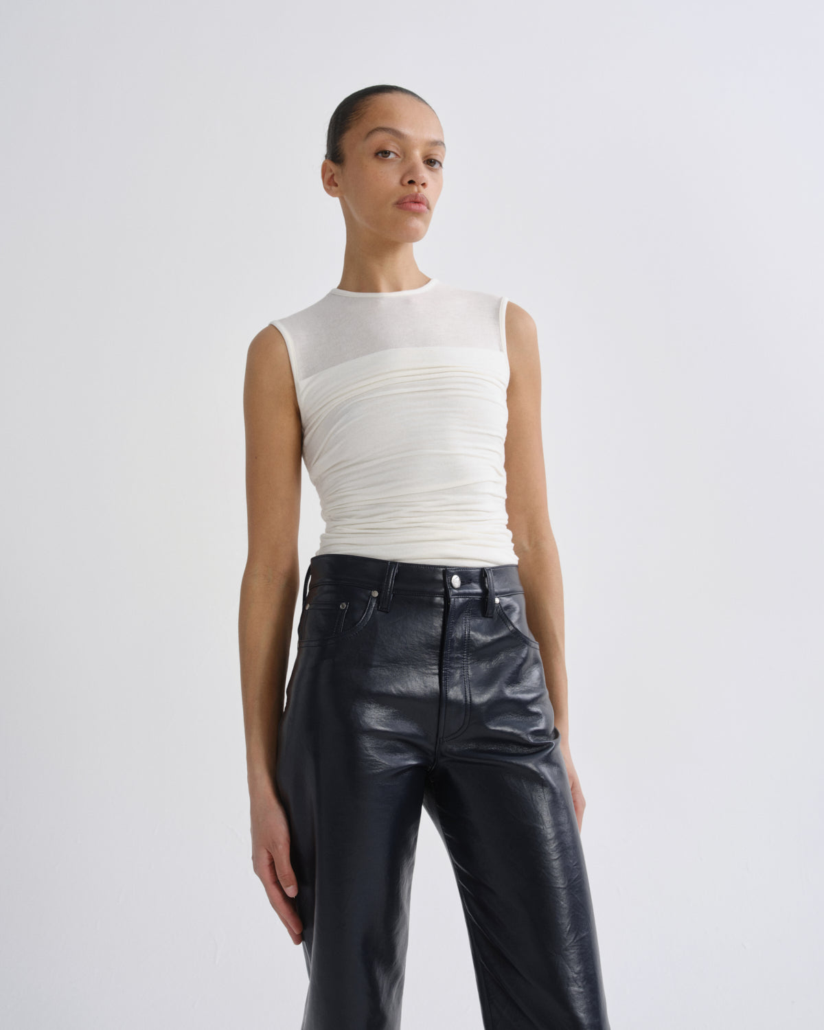 A model wearing a white sleeveless mesh and ruched top paired with black leather pants, standing against a light gray studio background.