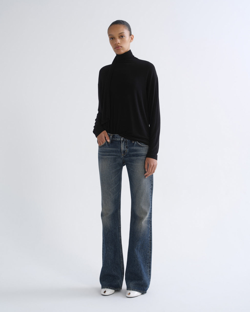 A model stands against a light gray studio background wearing a black long-sleeve turtleneck sweater, blue wide-leg jeans, and white pointed shoes.