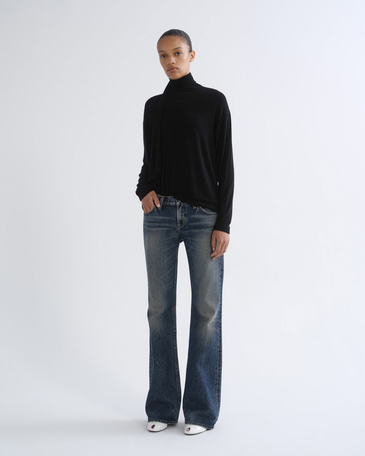 A model stands against a light gray studio background wearing a black long-sleeve turtleneck sweater, blue wide-leg jeans, and white pointed shoes.