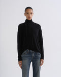 A model wearing a black long-sleeve turtleneck sweater with a draped tie detail at the neck, paired with blue jeans, against a plain light gray studio background.