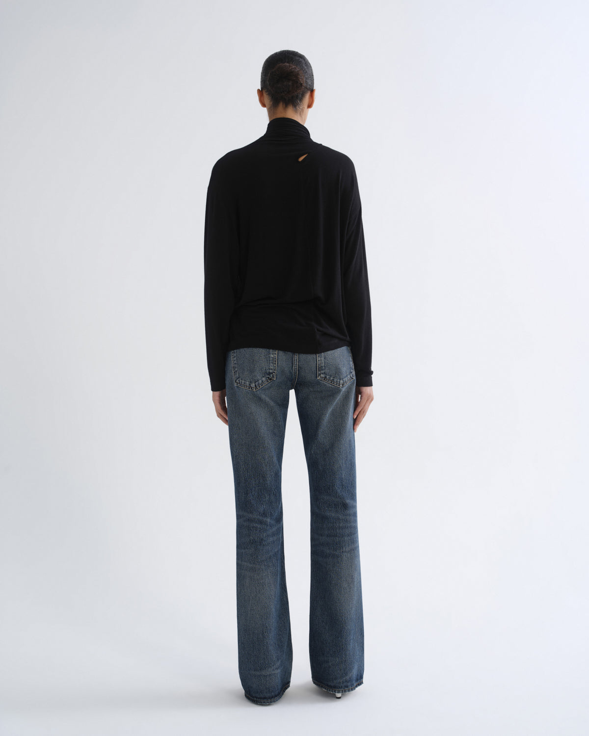A model viewed from the back wearing a black long-sleeve mock neck top and blue jeans against a plain light gray studio background.