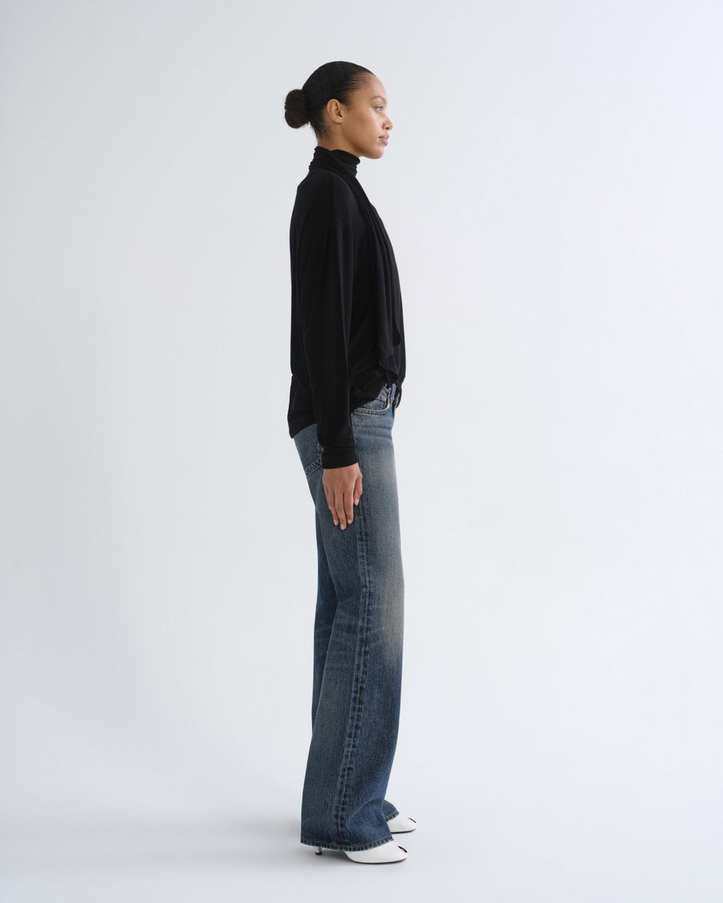 A model shown in profile wearing a black long-sleeve turtleneck top, blue wide-leg jeans, and white pointed shoes against a plain light gray studio background.