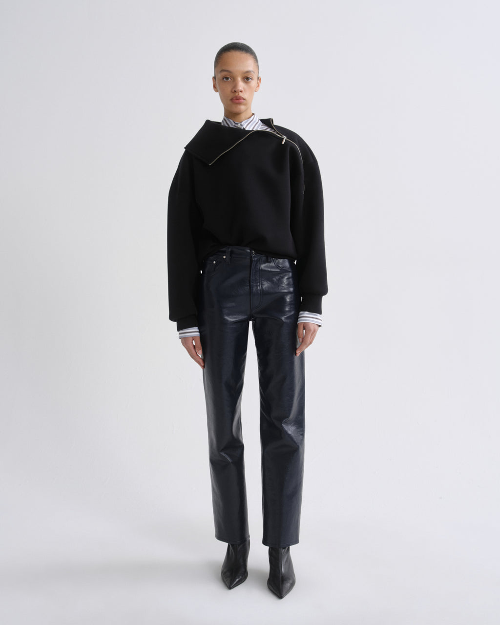 A model stands against a light gray studio background wearing a black asymmetrical knit sweater, shiny dark navy trousers, a striped shirt at the collar, and black pointed boots.