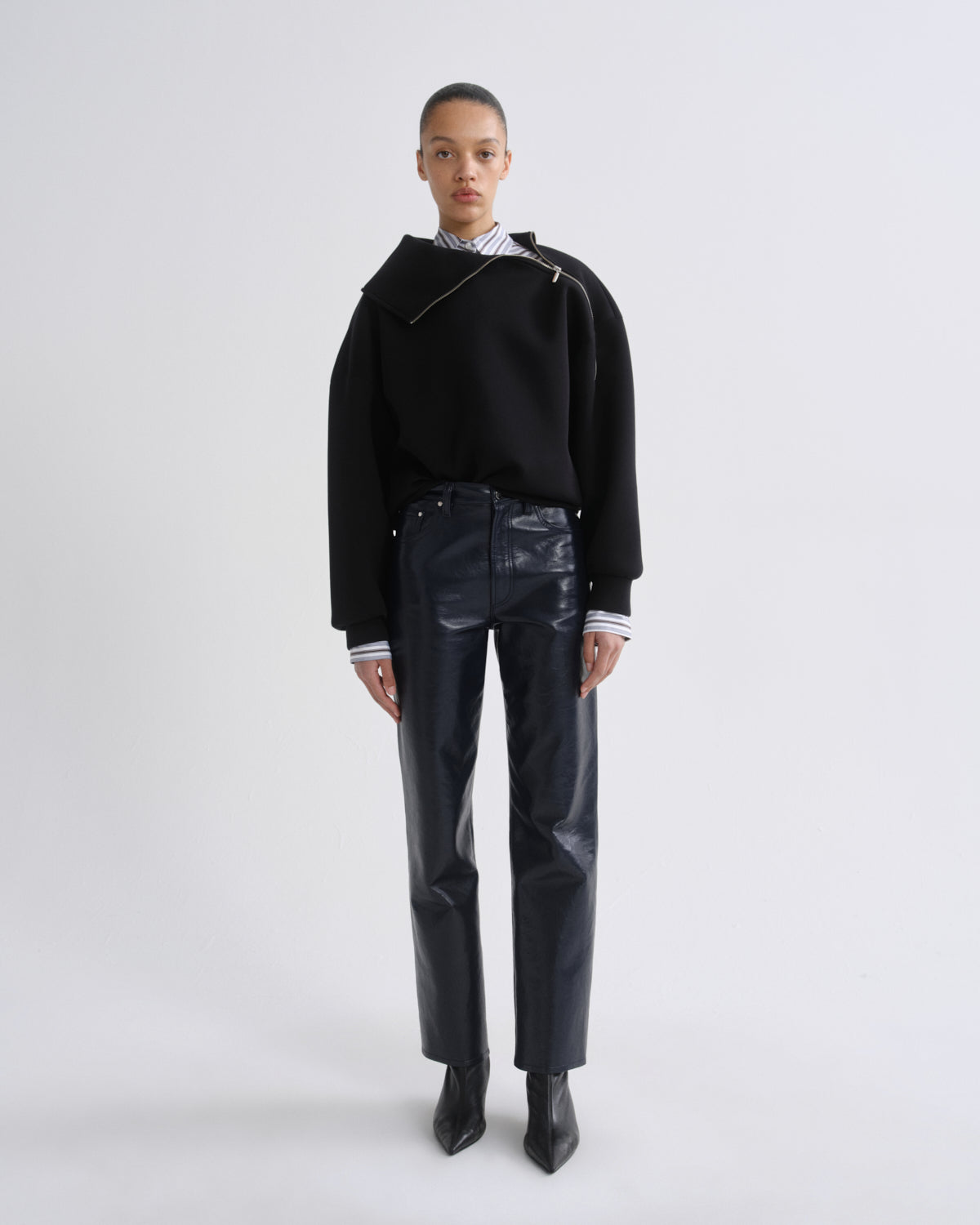 A model stands against a light gray studio background wearing a black asymmetrical knit sweater, shiny dark navy trousers, a striped shirt at the collar, and black pointed boots.