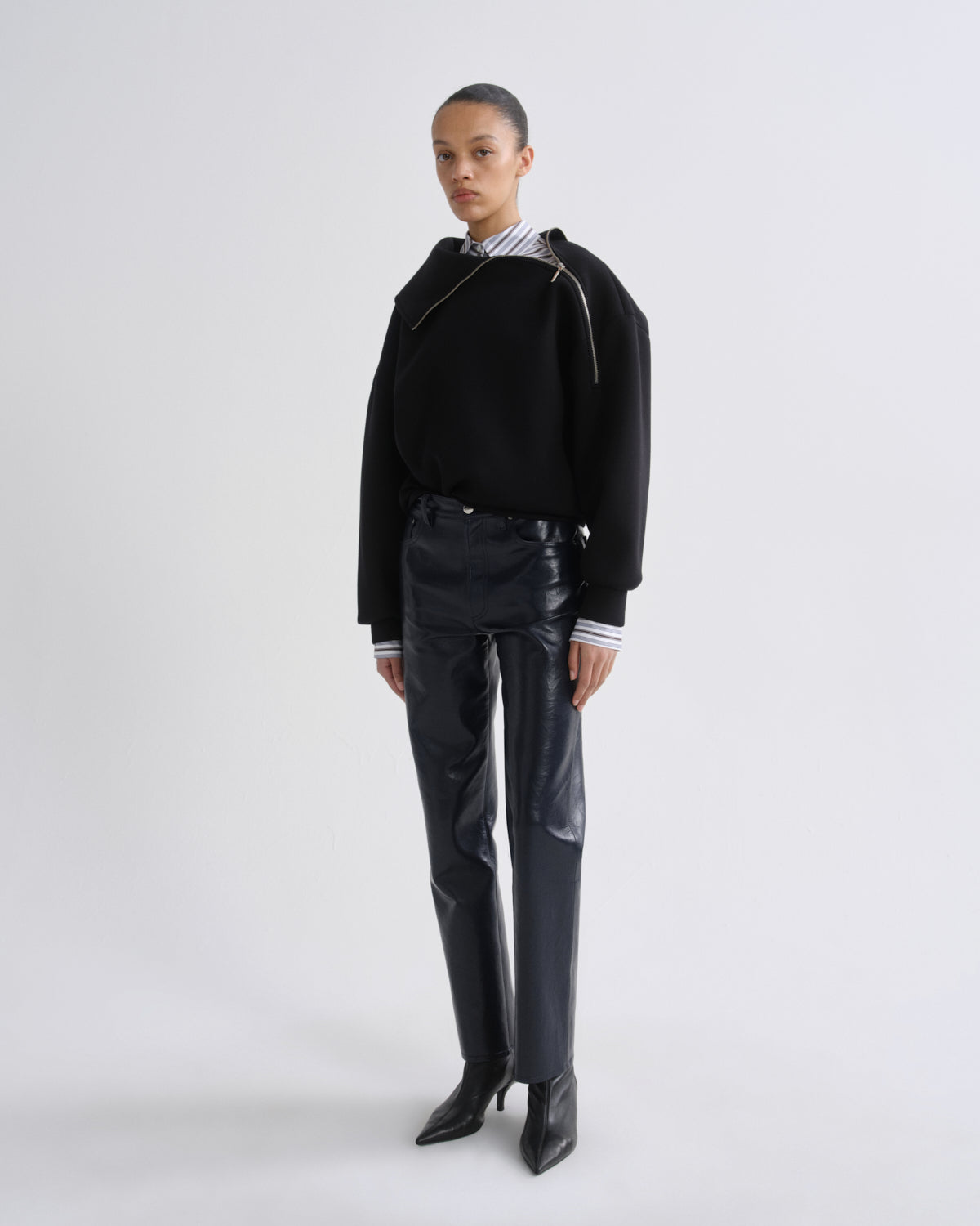 A model stands against a light gray studio background wearing a black asymmetrical zip-front sweatshirt over a striped shirt, paired with shiny black pants and black pointed ankle boots.