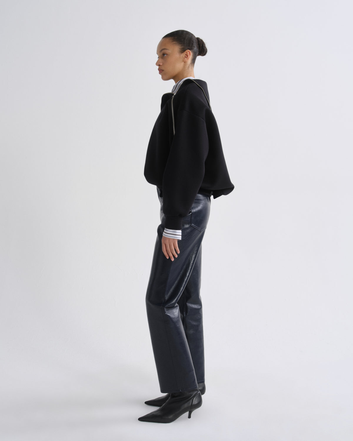 A model is shown in profile wearing a black cropped zip-up hoodie, shiny dark gray pants, and black pointed-toe heeled boots against a plain light gray studio background.