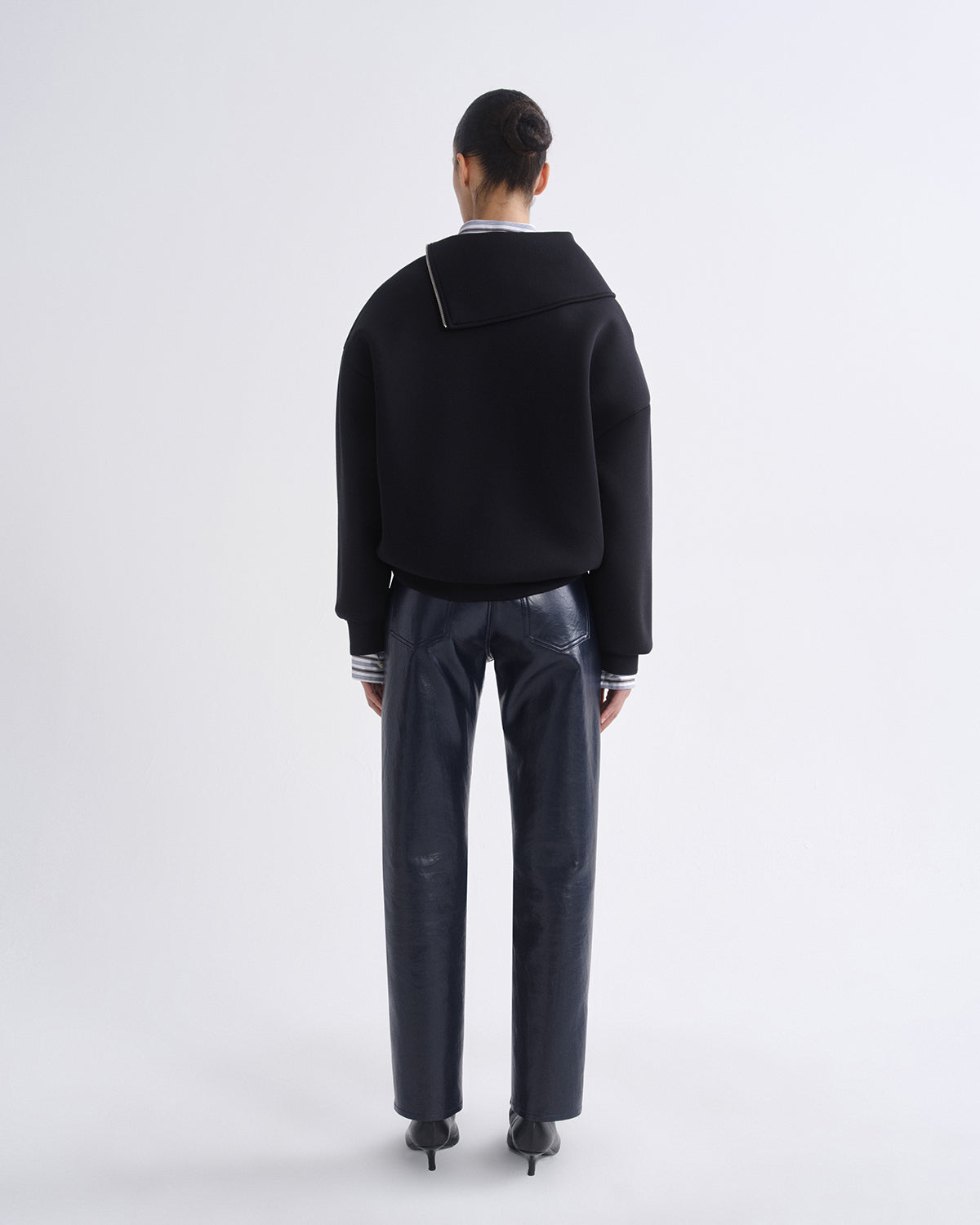 A model viewed from the back wearing a black cropped sweater with a wide collar and striped cuffs, paired with dark wide-leg trousers and black heels, against a light gray studio background.