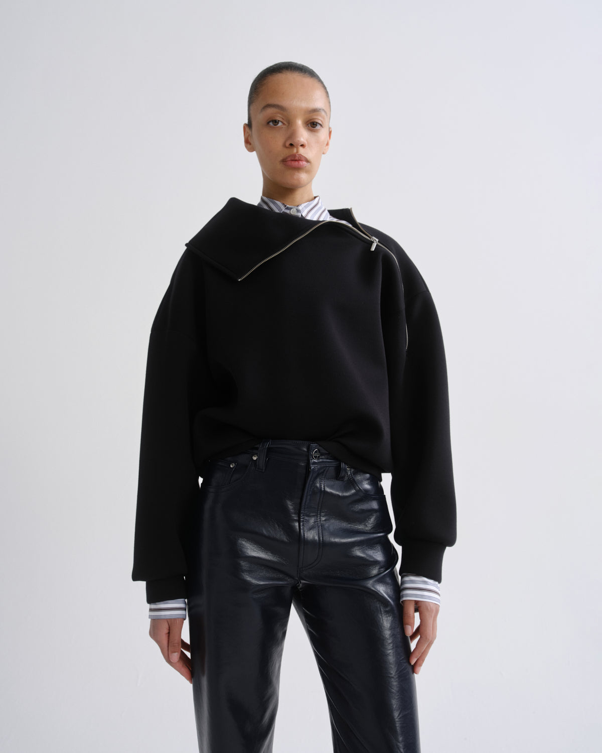 A model wearing a black asymmetrical zip sweatshirt with a wide collar, paired with black leather pants and a striped shirt, shot against a light gray studio background.