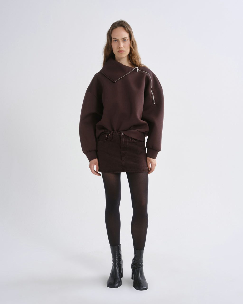 A model stands against a light gray studio background wearing a dark brown asymmetrical zip-up sweatshirt, a matching dark brown mini skirt, black tights, and black ankle boots.