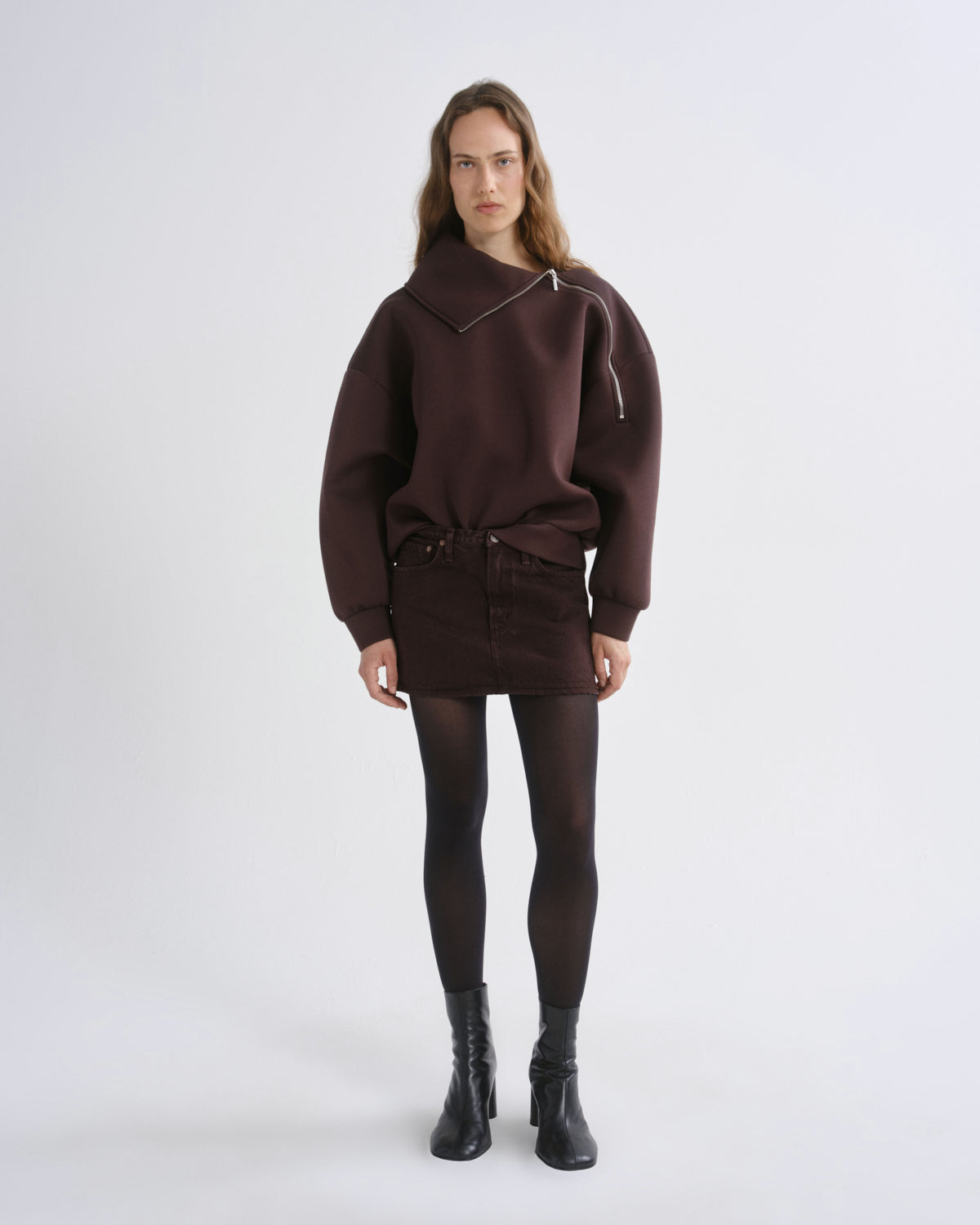 A model stands against a light gray studio background wearing a dark brown asymmetrical zip-up sweatshirt, a matching dark brown mini skirt, black tights, and black ankle boots.