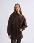 A woman modeling a dark brown oversized sweatshirt with a wide collar and silver zipper detail, paired with a matching dark brown mini skirt, against a light gray studio background.