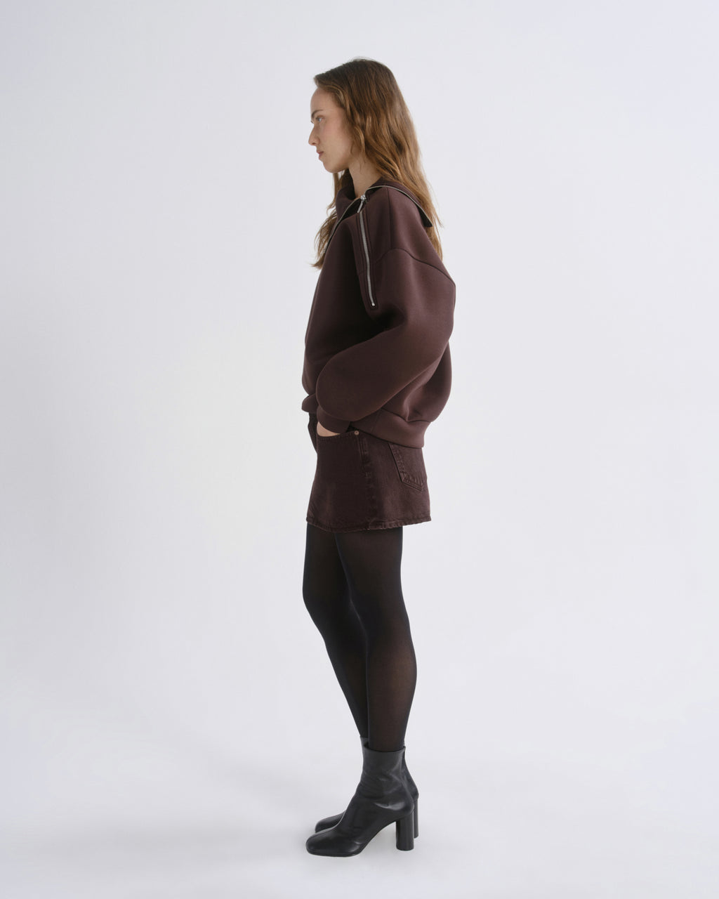 A woman stands in profile wearing a dark brown hooded sweatshirt, a matching brown mini skirt, sheer black tights, and black ankle boots against a plain light gray studio background.