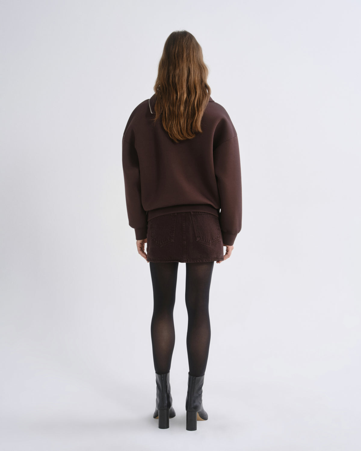 A model viewed from the back wearing a dark brown knit sweater and matching mini skirt, paired with black tights and black ankle boots, against a light gray studio background.
