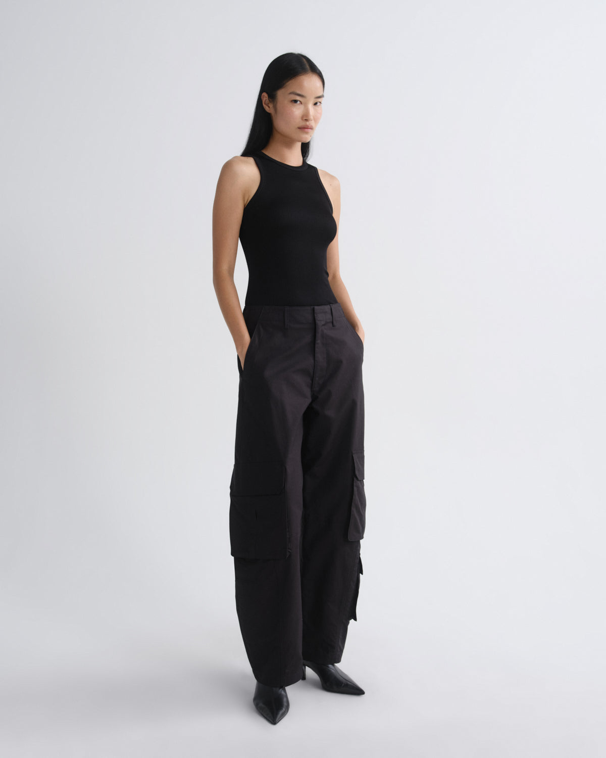 A model stands in a studio wearing a black sleeveless top and black wide-leg cargo pants with side pockets, paired with black pointed shoes, against a light gray background.