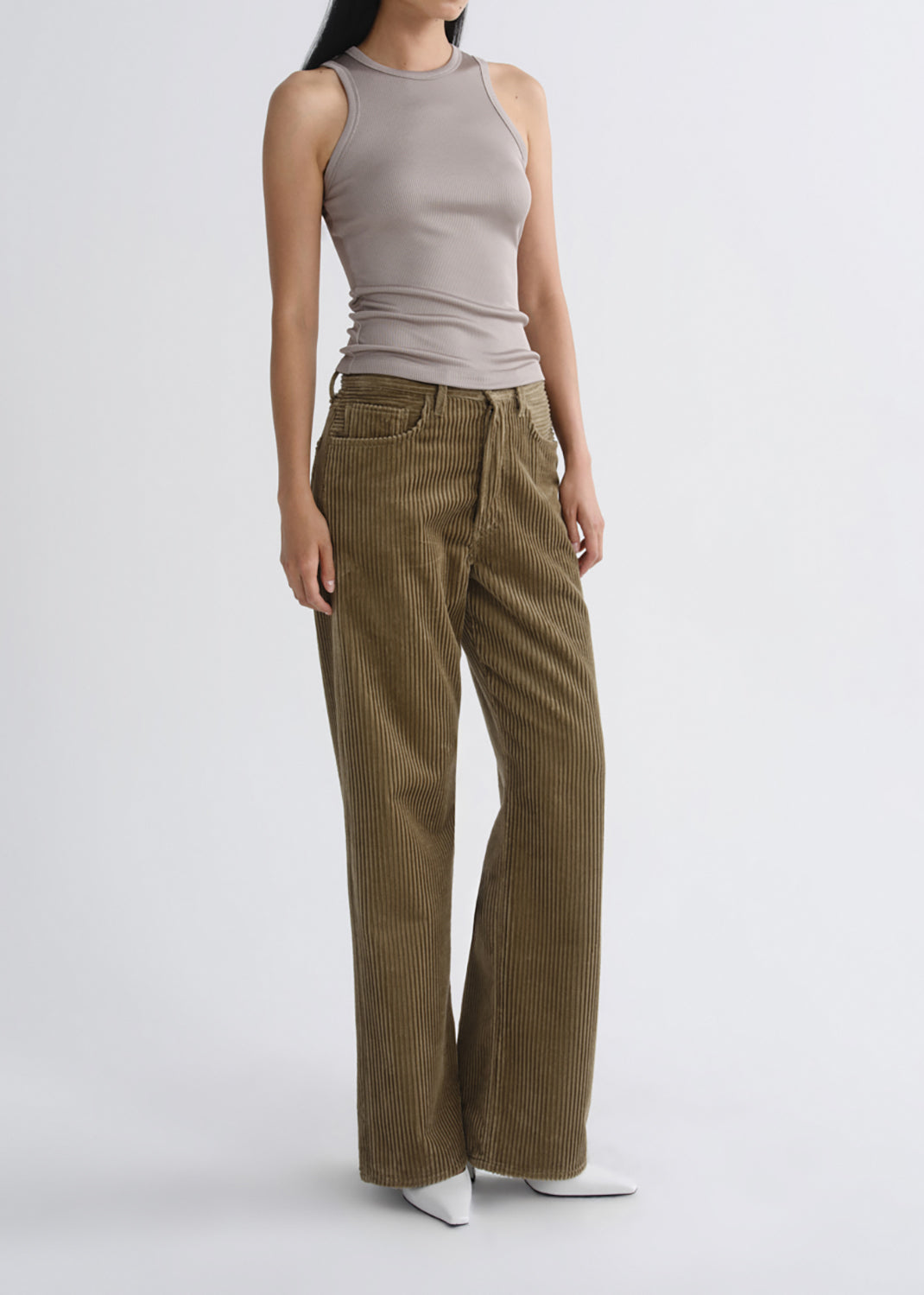 A model wearing olive brown corduroy wide-leg pants, a taupe sleeveless ribbed top, and white pointed-toe heels, standing against a grey background.