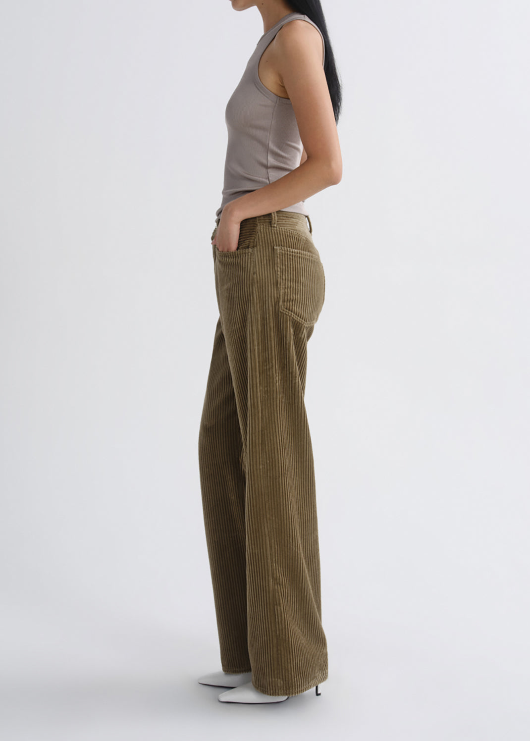 A model shown from the side wearing high-waisted, wide-leg corduroy pants in an olive-brown color, paired with a taupe tank top and white pointed-toe heels, against a grey background.
