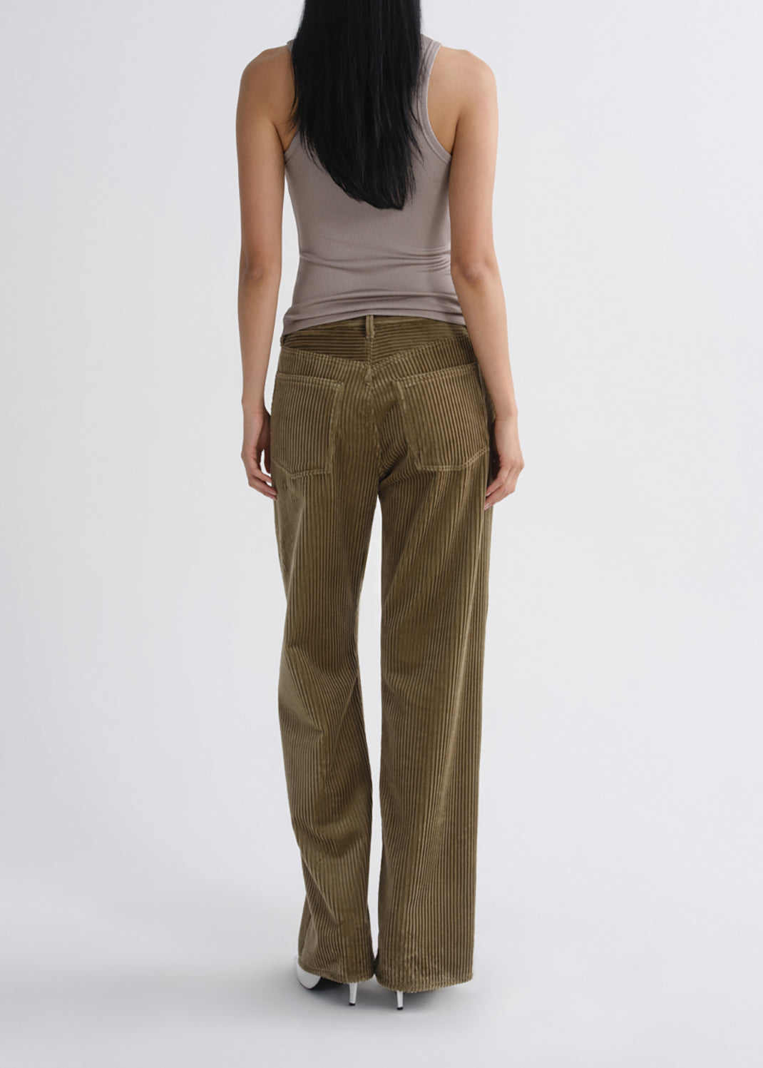 A woman viewed from the back wearing olive brown corduroy wide-leg pants, a taupe tank top, and white heels against a grey background.