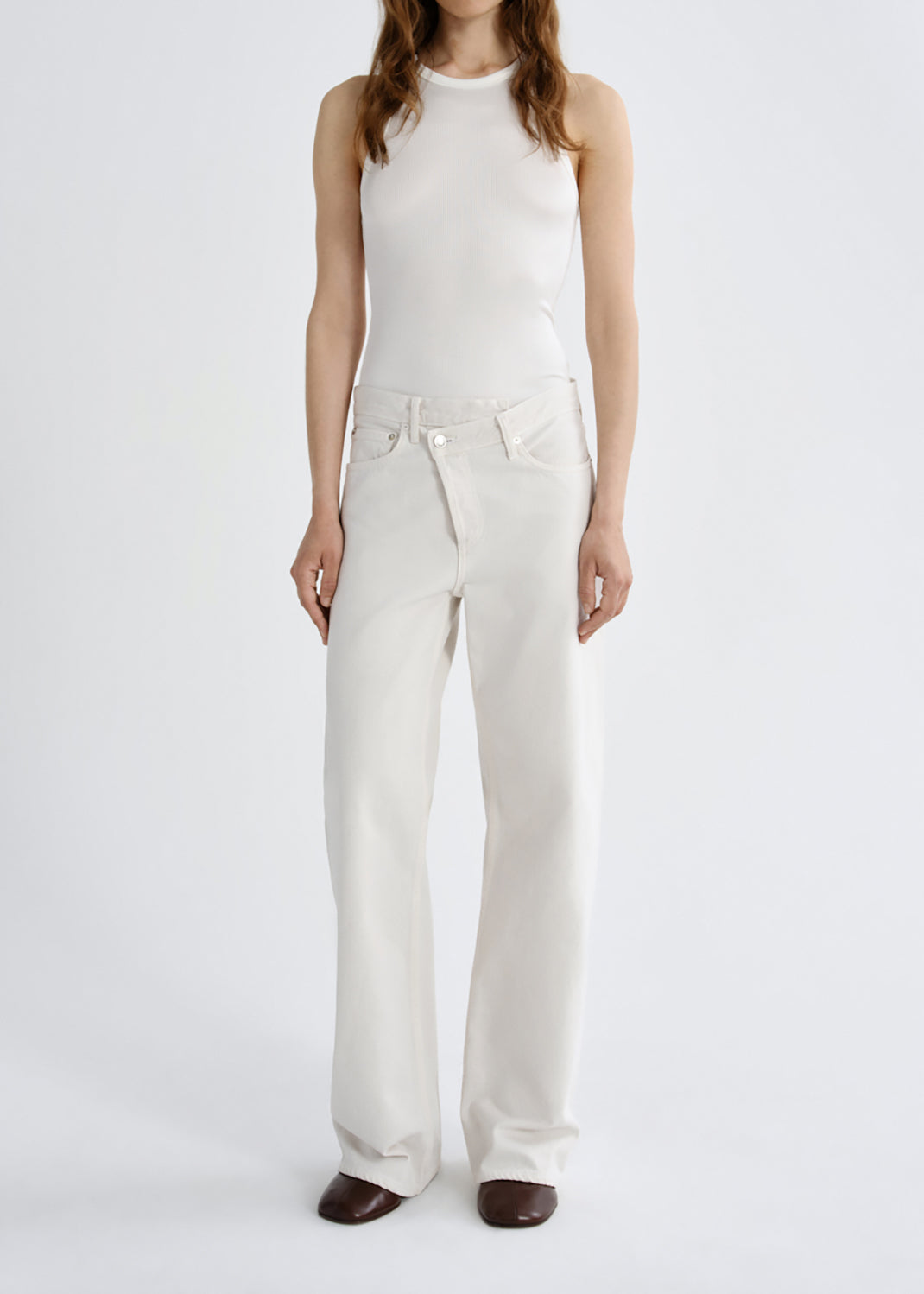 A model wearing a white sleeveless mock-neck top and matching white wide-leg trousers, paired with dark brown pointed-toe flats, against a grey background.