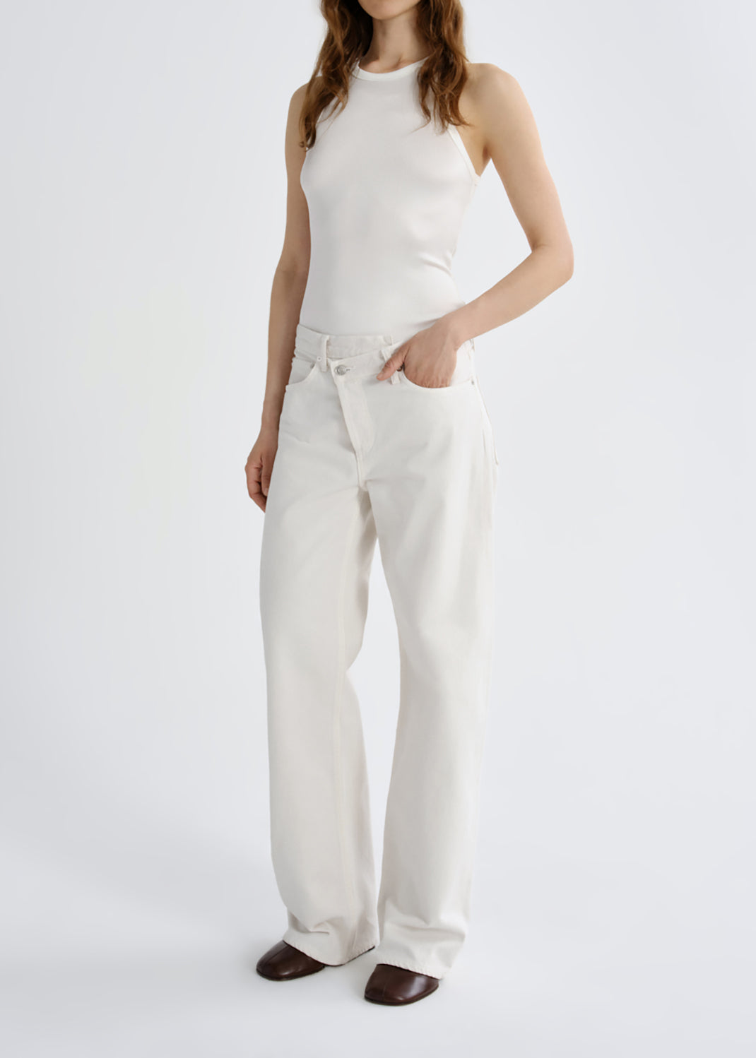 A model wearing a white sleeveless mock-neck top and matching white wide-leg trousers, paired with dark brown pointed-toe flats, against a grey background.