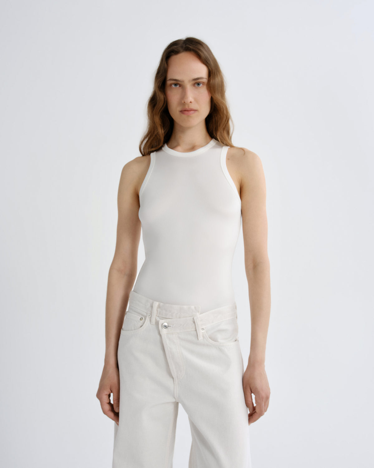 Woman wearing a white sleeveless top and light-colored pants on a plain background