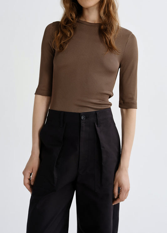 A woman wearing a fitted brown short-sleeve top and black high-waisted trousers, photographed against a clean grey background.