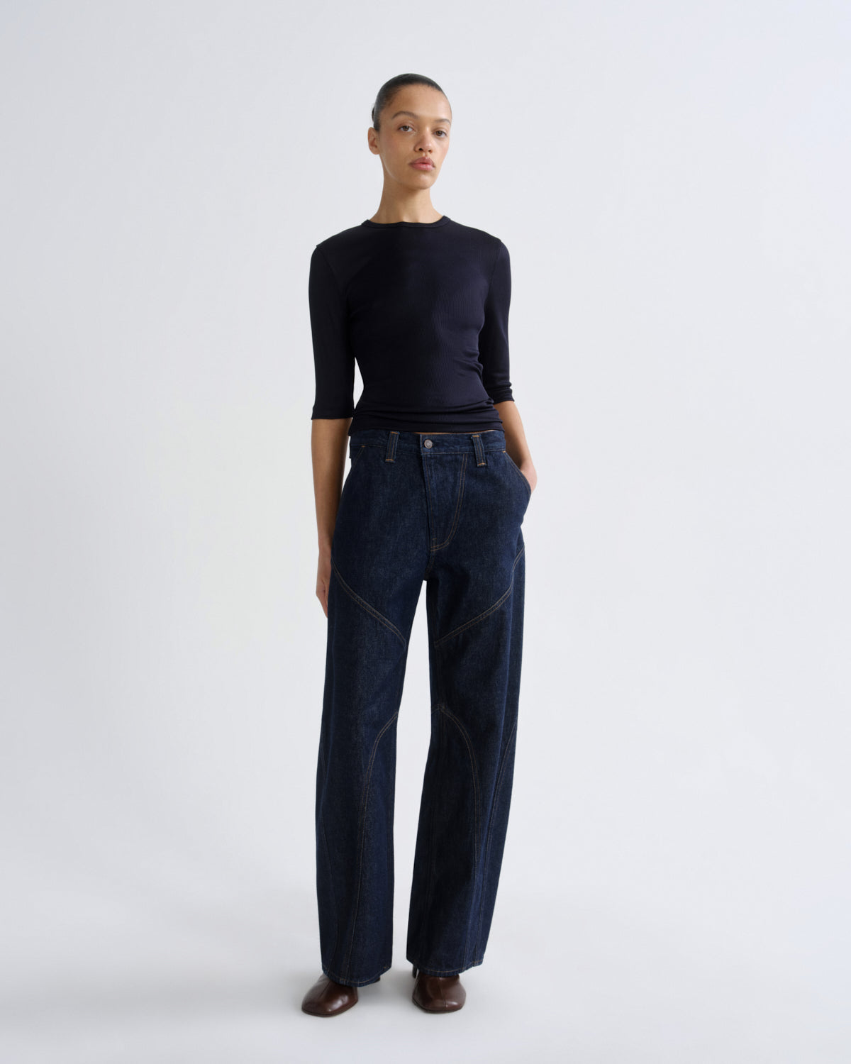 A model stands against a light gray studio background wearing a dark navy long-sleeve top and dark blue wide-leg jeans with brown shoes.