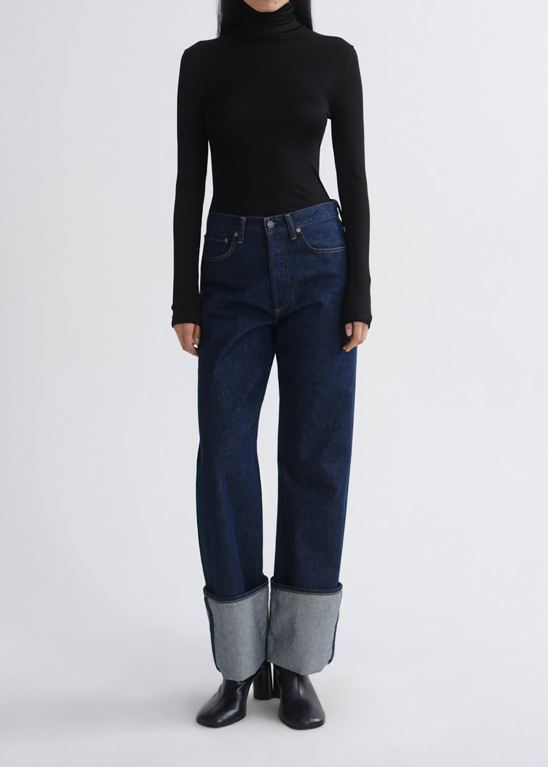 A model wearing dark blue straight-leg jeans with a rolled-up hem showing light blue denim lining, paired with a black long-sleeve turtleneck and black boots, against a grey background.