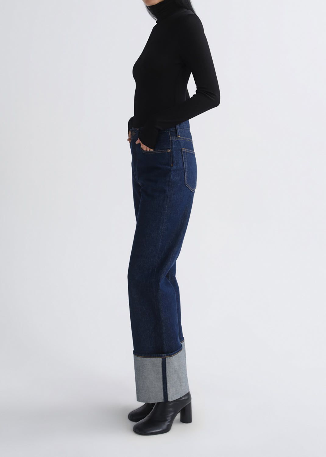 A model shown from the side wearing dark blue cuffed jeans, a black long-sleeve turtleneck, and black heeled boots against a grey background.