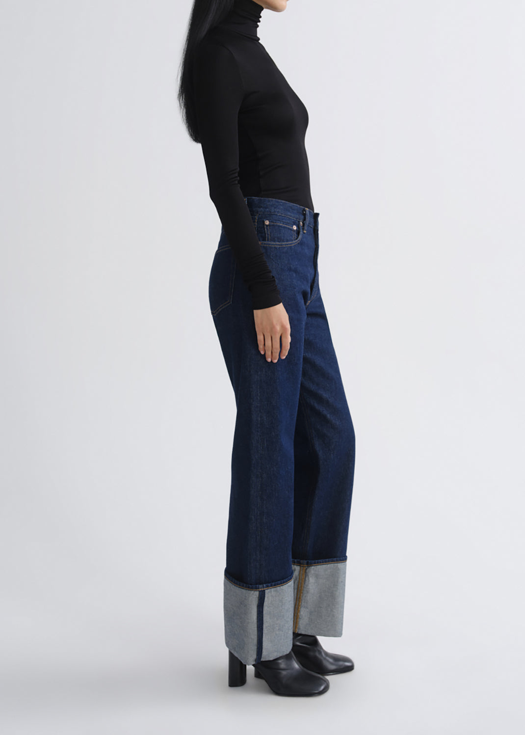 A model shown from the side wearing dark blue wide-leg jeans with a contrasting light gray inner cuff, a black long-sleeve top, and black ankle boots, against a grey background.
