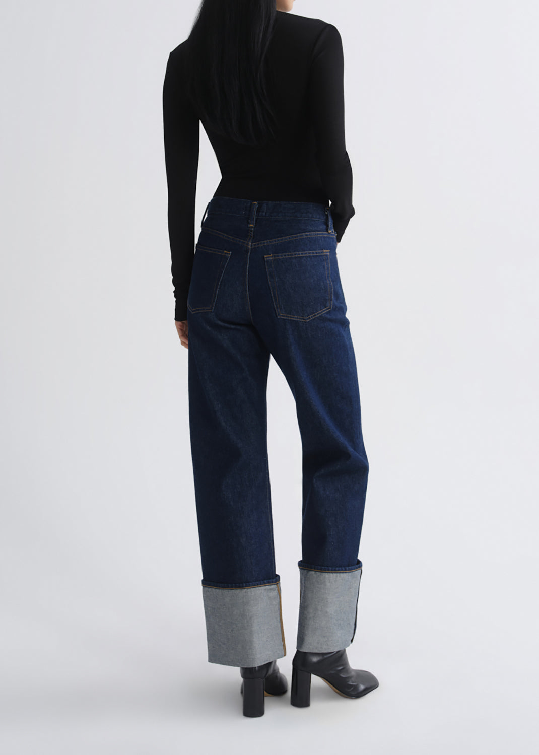 A model shown from the back wearing dark blue straight-leg jeans with rolled cuffs, a black long-sleeve top, and black boots, against a grey background.