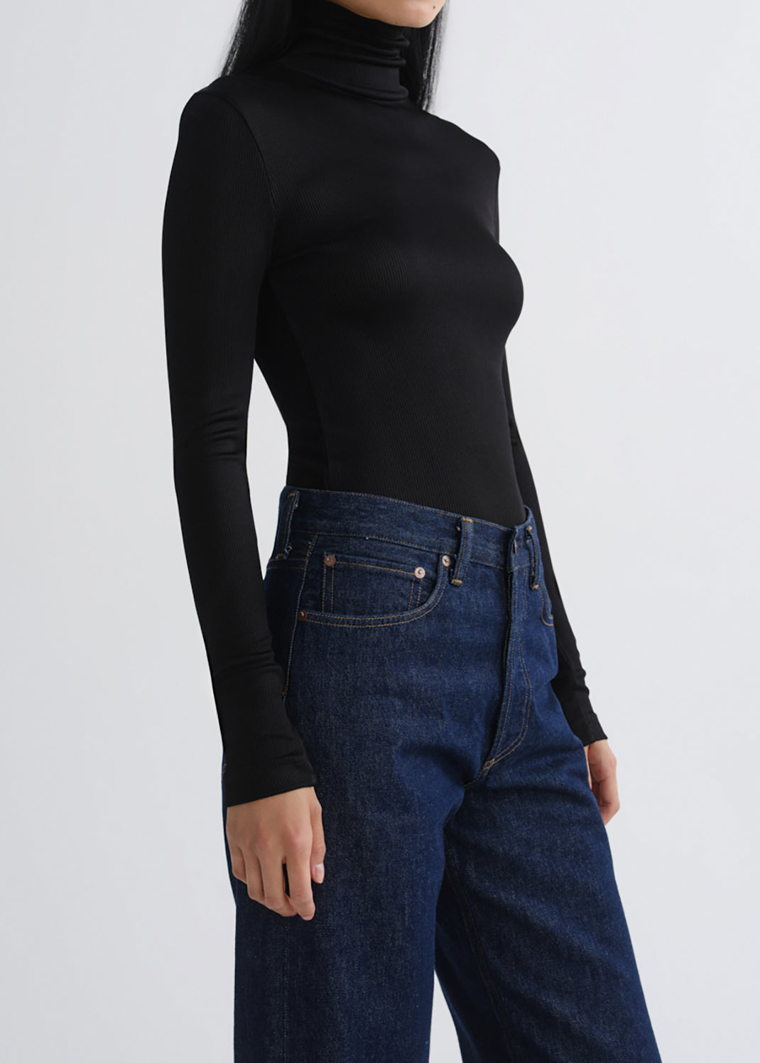 A model wearing a fitted black long-sleeve turtleneck top and dark blue jeans, shown from the side against a grey background.