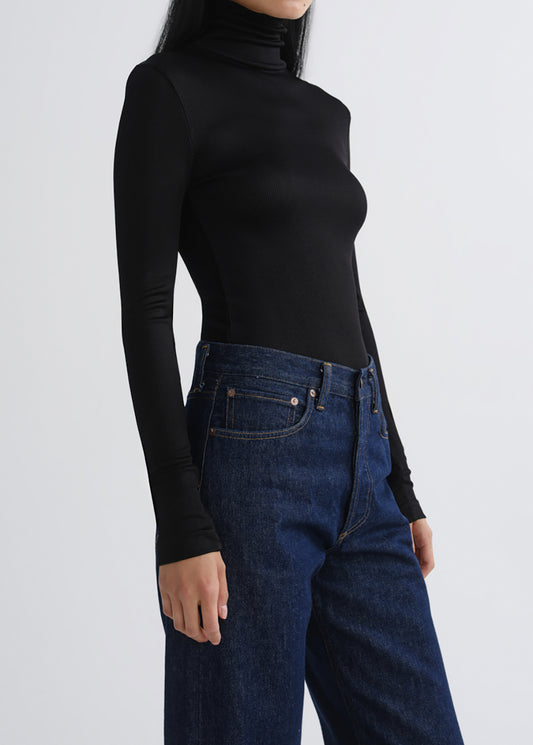 A model wearing a fitted black long-sleeve turtleneck top and dark blue jeans, shown from the side against a grey background.