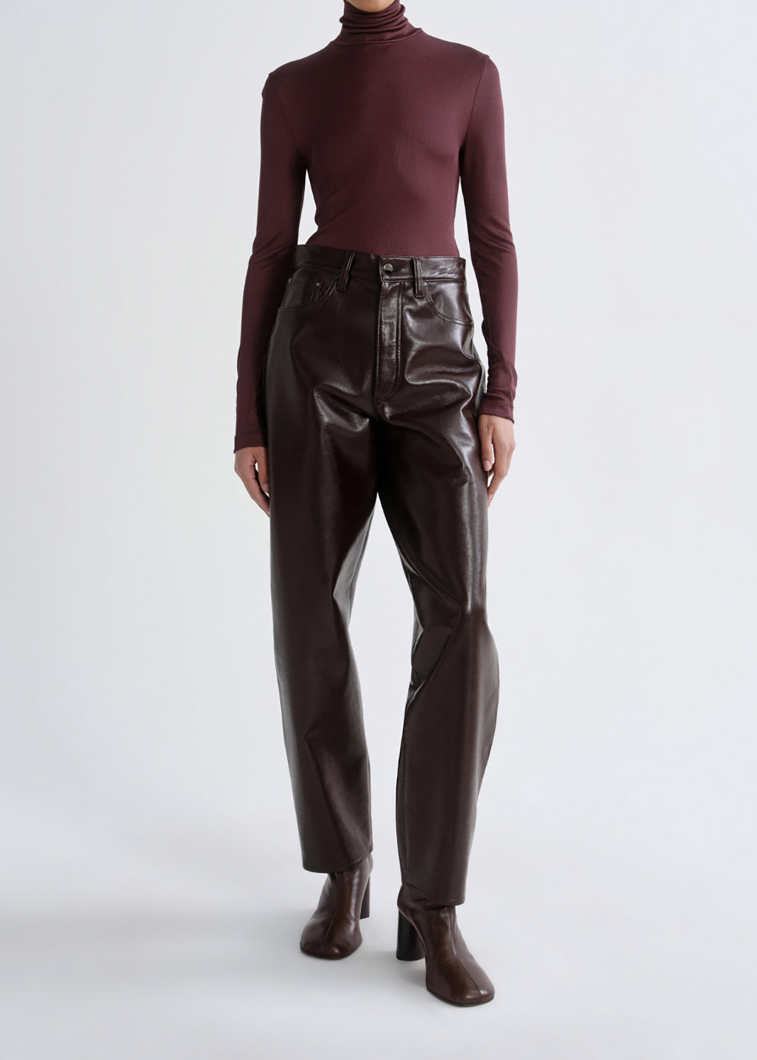 A model wearing a dark burgundy ribbed turtleneck top and high-waisted, glossy dark brown faux-leather trousers with dark brown block-heel boots, photographed against a grey background.