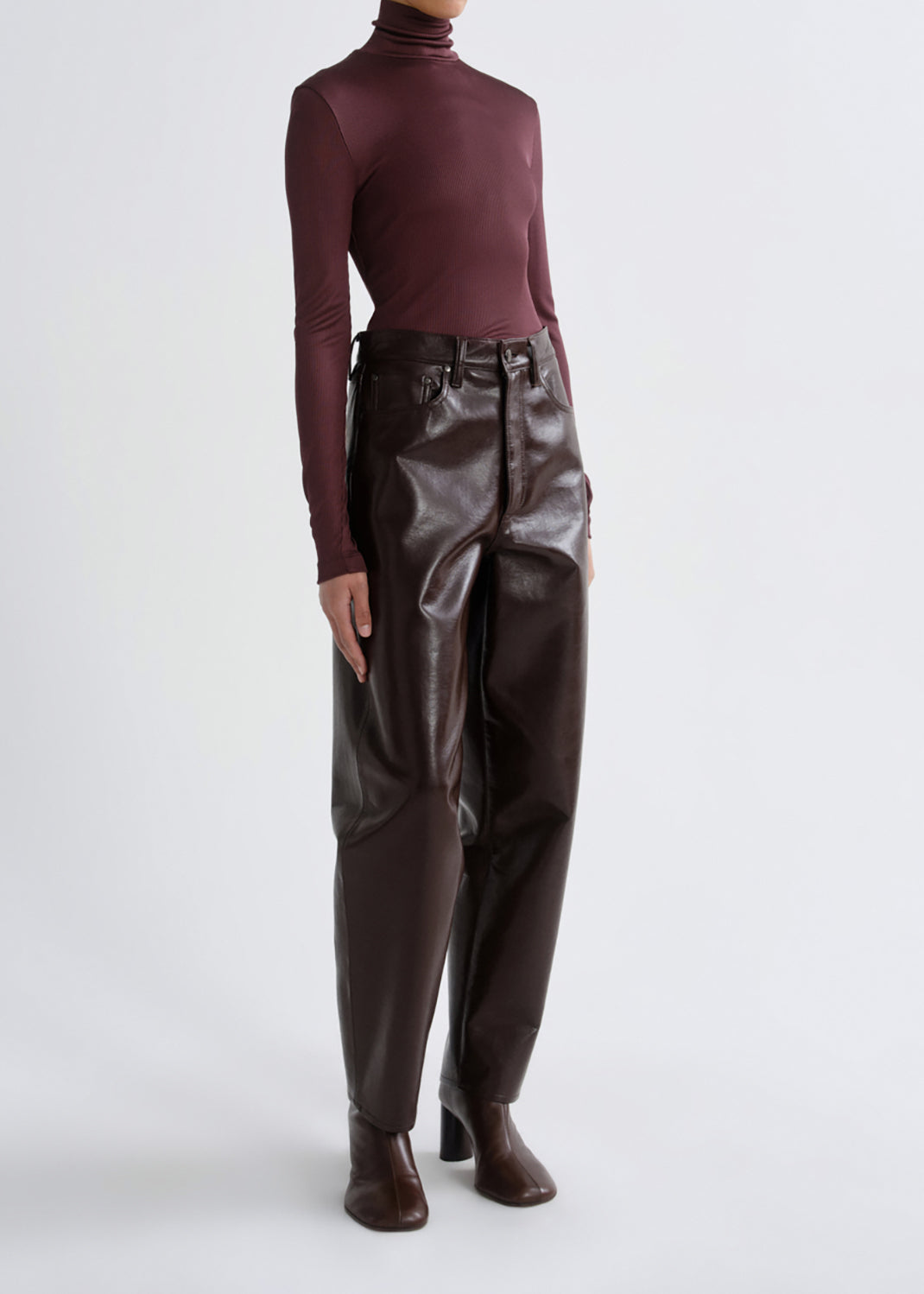 A model wearing a dark burgundy long-sleeve turtleneck top and dark brown faux-leather trousers with a matching belt, paired with dark brown heeled ankle boots, against a grey background.