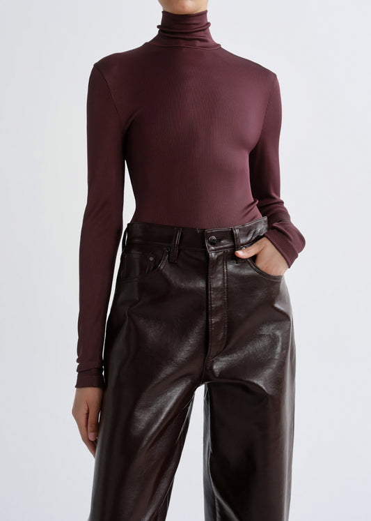 A model wearing a fitted long-sleeve turtleneck top in deep burgundy and high-waisted dark brown faux-leather pants, shown from the neck down against a grey background.