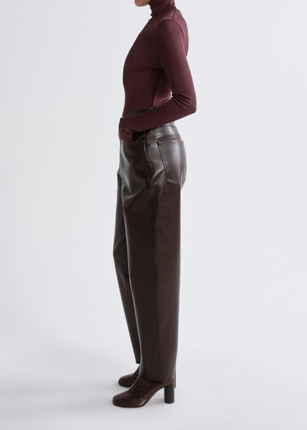 A model shown from the side wearing dark brown leather trousers, a burgundy turtleneck top, and dark brown heeled boots against a grey background.