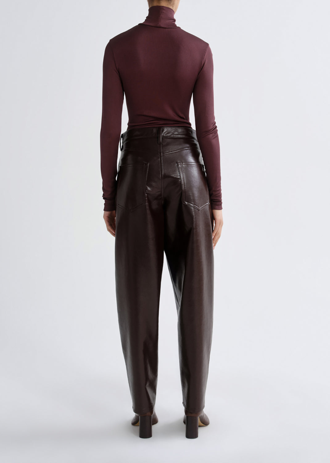 A model shown from the back wearing a dark burgundy long-sleeve mock neck top and high-waisted dark brown faux-leather trousers, set against a grey background.