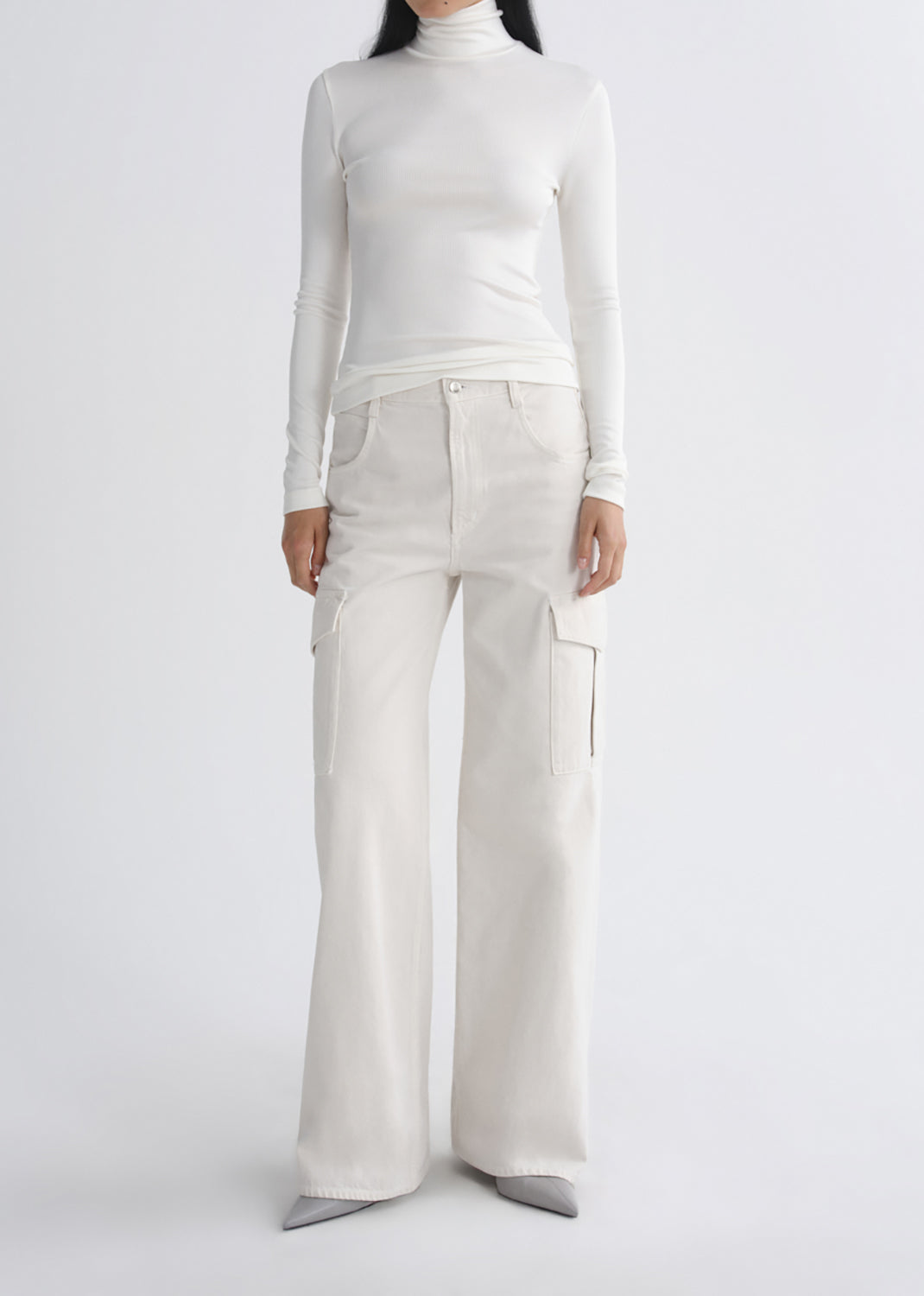 A model wearing a white long-sleeve mock neck top and matching off-white wide-leg cargo pants, paired with grey pointed shoes, against a grey background.