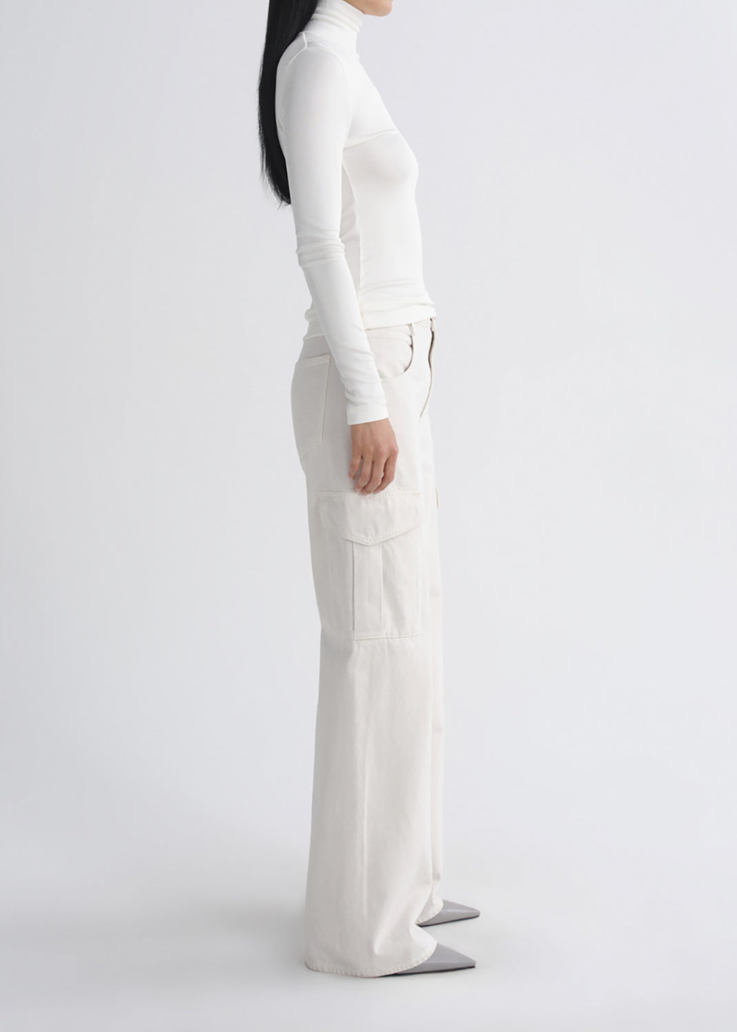 A model shown from the side wearing a white long-sleeve turtleneck top and matching white wide-leg cargo pants, paired with grey pointed-toe flats, against a grey background.