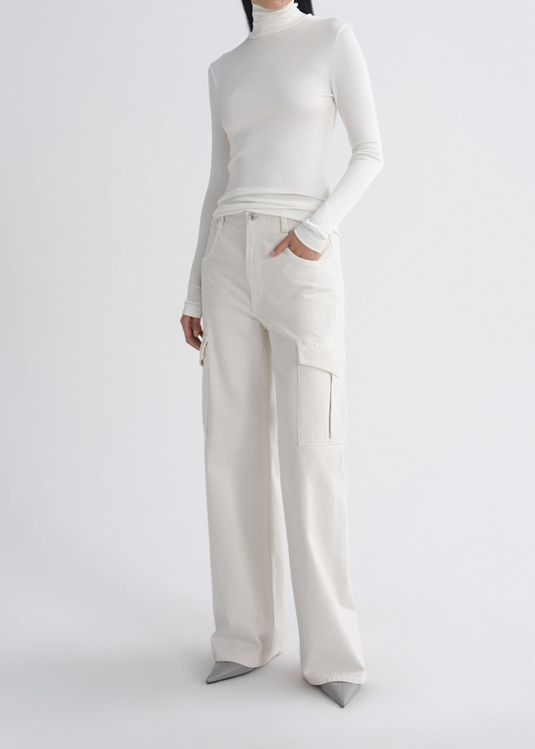 A model wearing a white long-sleeve mock-neck top and off-white wide-leg cargo pants with grey pointed-toe heels, photographed against a grey background.