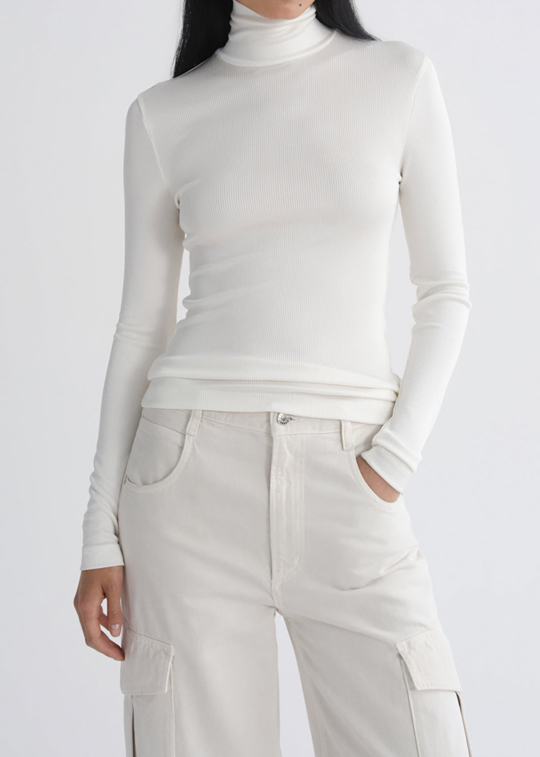 A model wearing a fitted, long-sleeve white turtleneck top and matching off-white cargo pants, shown from the neck down against a grey background.