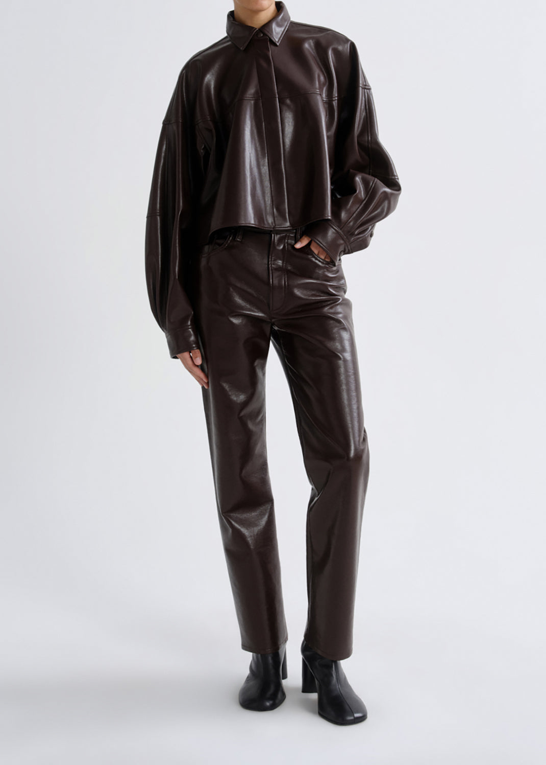 A model wearing a matching dark brown faux leather two-piece set consisting of a long-sleeve collared shirt and straight-leg trousers, paired with black ankle boots, against a grey background.