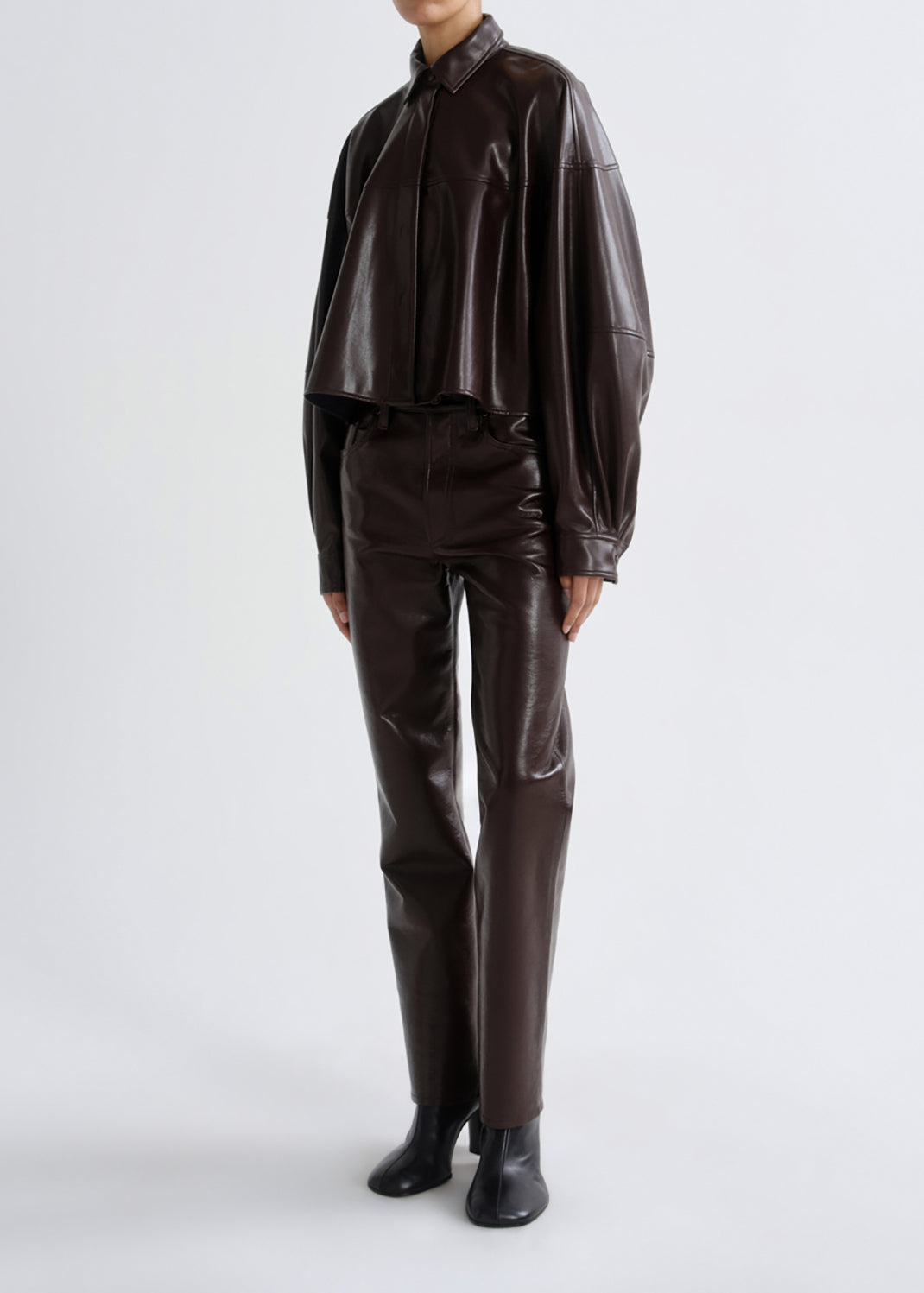 A model wearing a dark brown faux leather two-piece set consisting of a cropped long-sleeve button-up shirt and matching slim-fit trousers, paired with black ankle boots, shot against a grey background.