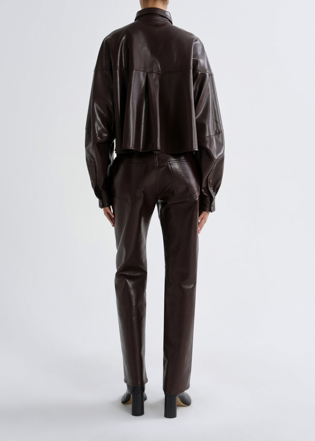 A model viewed from the back wearing a dark brown faux-leather cropped jacket and matching slim-fit trousers, paired with black heels, against a grey background.